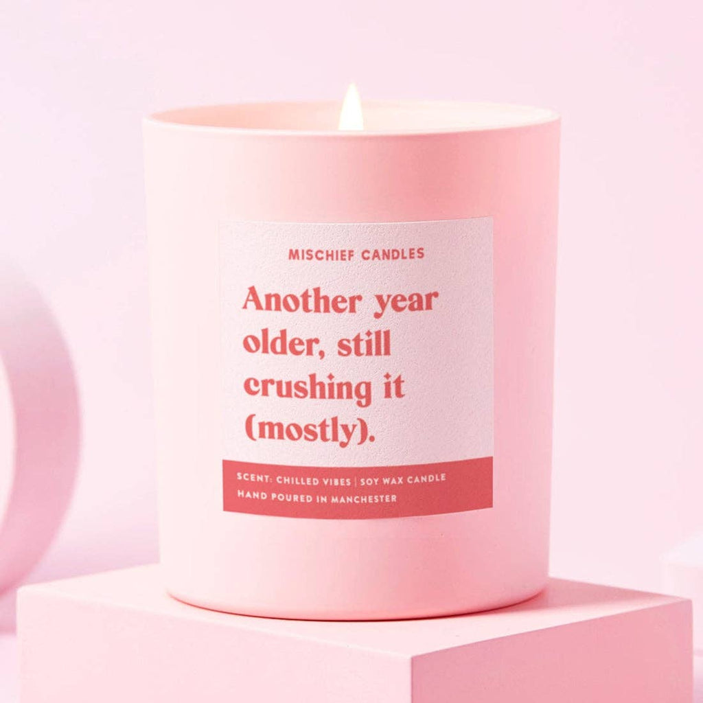 A pink jar scented candle with the text 'Another year older, still crushing it (mostly)' on a colorful label, placed on a pink surface.