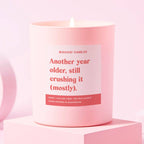 A pink jar scented candle with the text 'Another year older, still crushing it (mostly)' on a colorful label, placed on a pink surface.