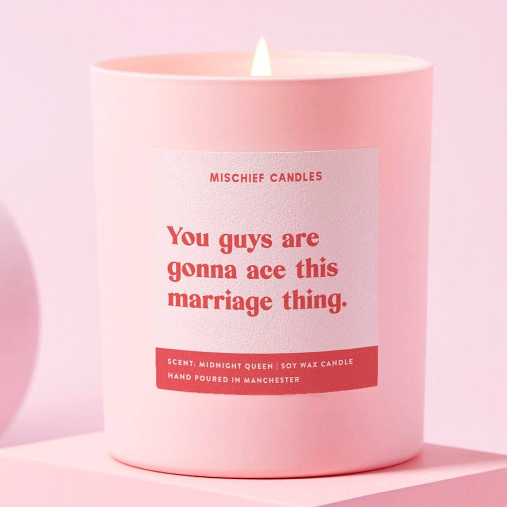 A pale pink candle with red and white text that reads 'You guys are gonna ace this marriage thing' with a lit flame on the top.