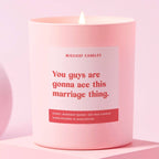 A pale pink candle with red and white text that reads 'You guys are gonna ace this marriage thing' with a lit flame on the top.