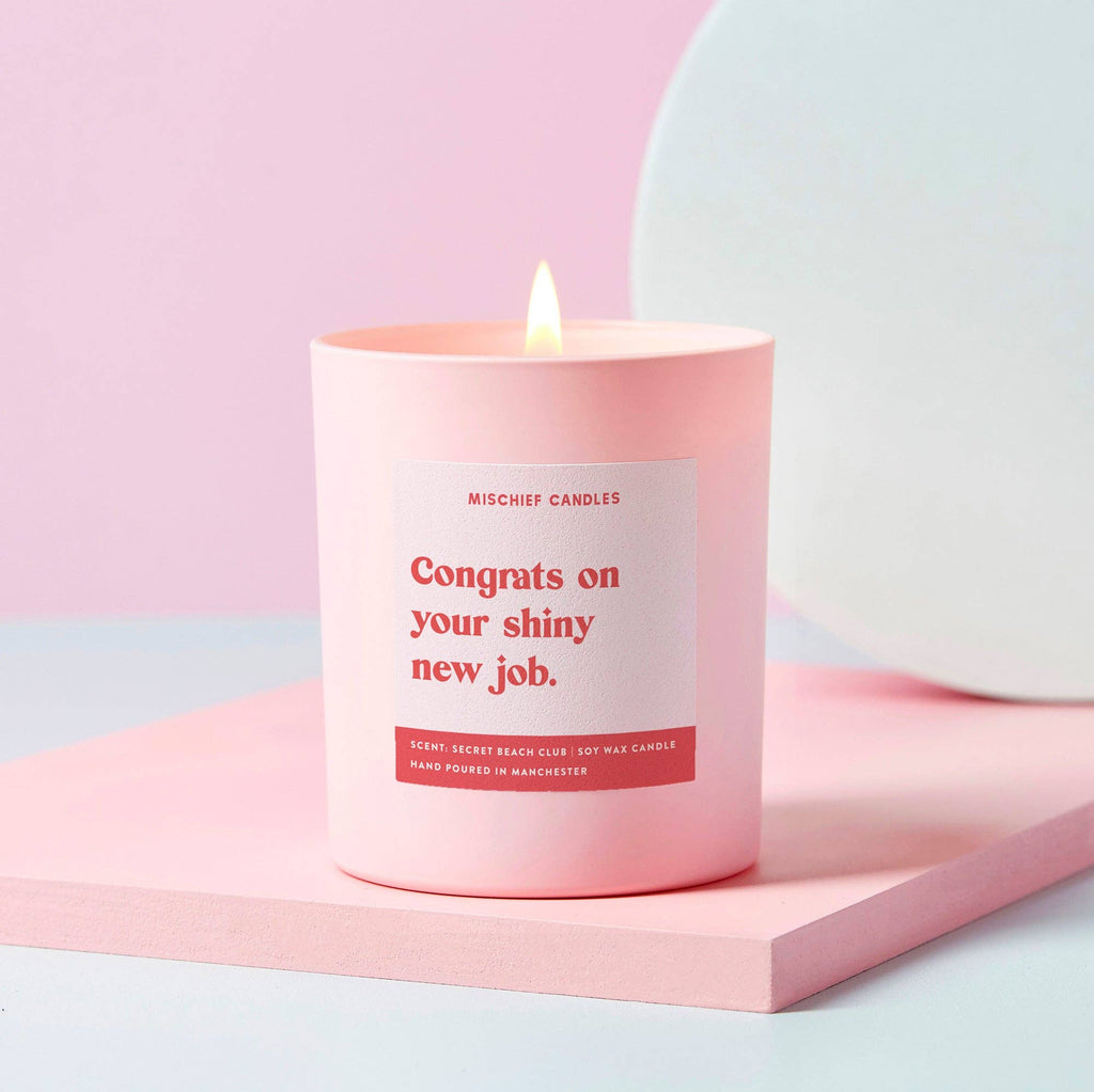 A pale pink candle with the text 'Congrats on your shiny new job' written in bold red font on the label. The candle is lit and placed on a pink surface with a white background.