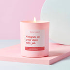 A pale pink candle with the text 'Congrats on your shiny new job' written in bold red font on the label. The candle is lit and placed on a pink surface with a white background.