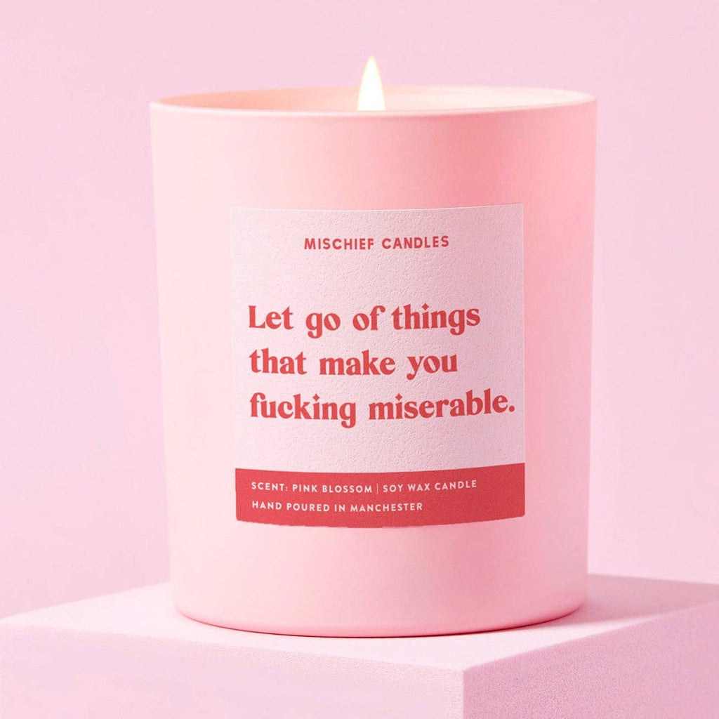 A pale pink candle with a red label that reads 'Let go of things that make you fucking miserable'. 