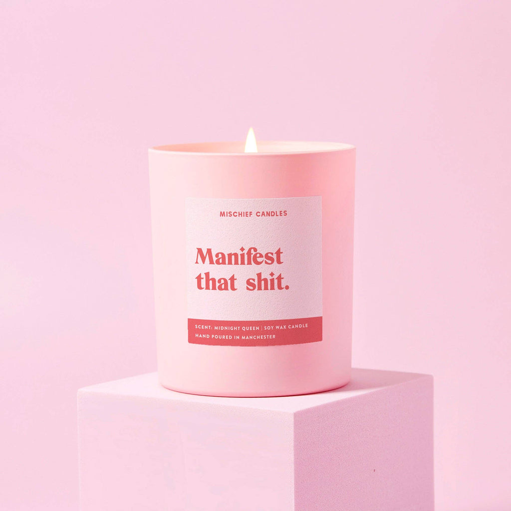 A pale pink candle with a label reading 'Manifest that shit' in bold red font, placed on a pink pedestal against a pink background.