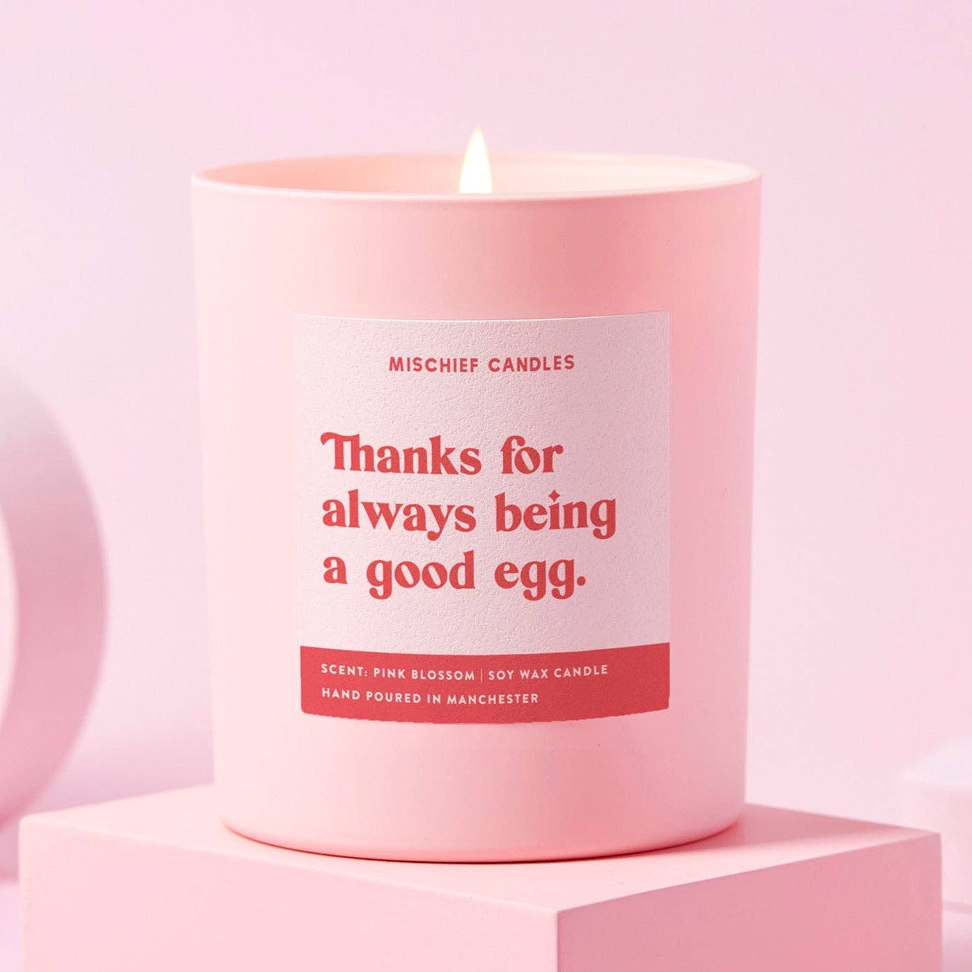 A pale pink candle with a red label reading 'Thanks for always being a good egg' with a lit flame, placed on a pink surface.