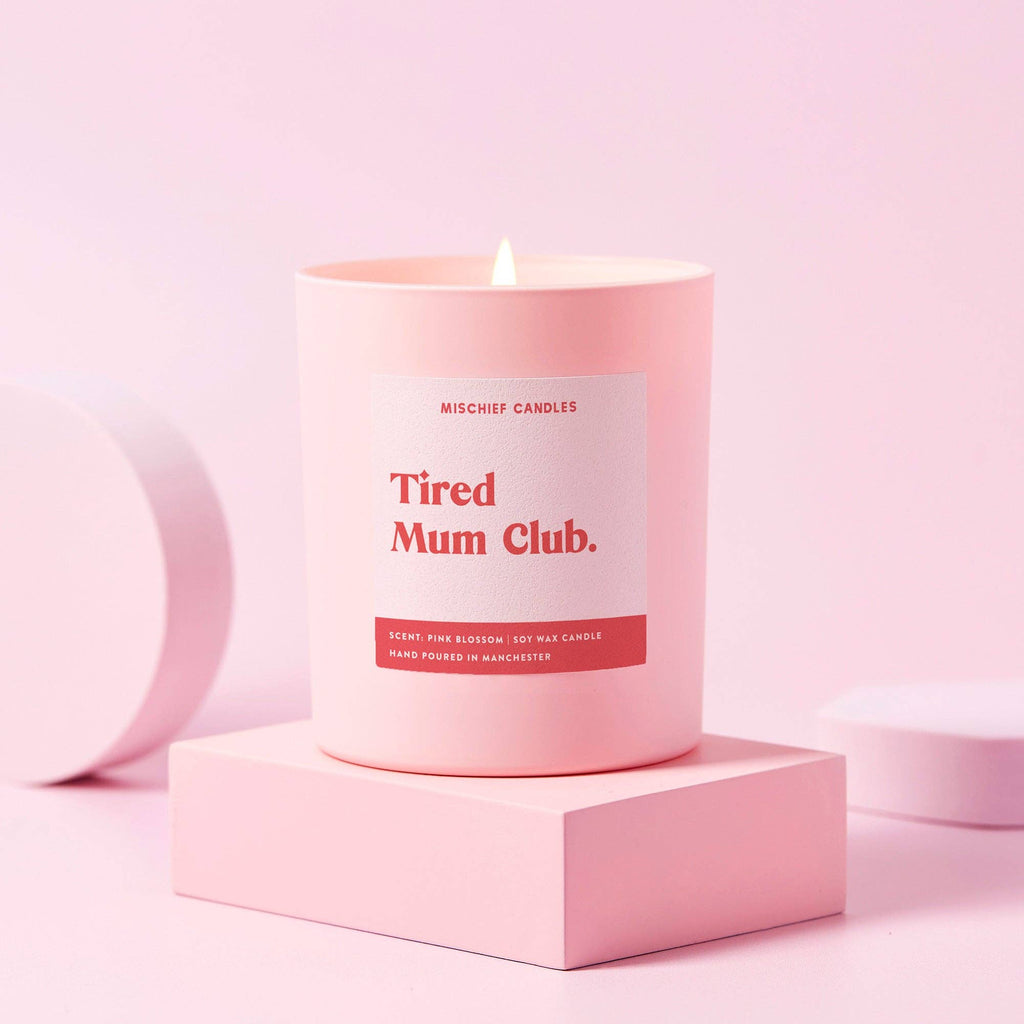 A pale pink candle with the text 'Tired Mum Club' in bold red font on the label, placed on a matching pink stand, against a pink background.