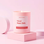 A pale pink candle with the text 'Tired Mum Club' in bold red font on the label, placed on a matching pink stand, against a pink background.
